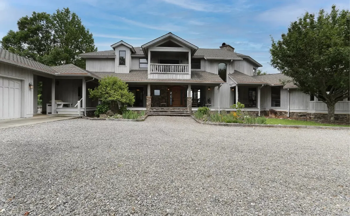 The image shows the main entrance of the estate, featuring impressive wooden double doors set against a backdrop of lush greenery and a gravel driveway. The porch area offers a welcoming transition between the outdoors and the home's interior, hinting at both privacy and hospitality.