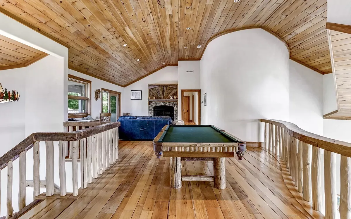This interior image highlights a welcoming game loft featuring a pool table set against a backdrop of large windows, a cozy fireplace, and a sturdy wooden railing. Natural light streams into the space, giving it a lively yet comfortable atmosphere ideal for both daytime fun and evening relaxation.