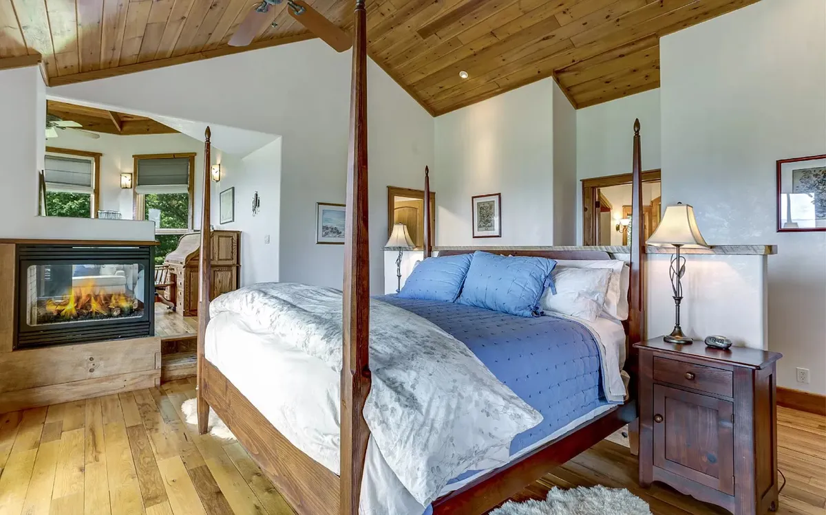 A beautifully appointed bedroom with a four-poster bed, a stone fireplace, warm wooden accents, and decorative lamps. The space feels inviting, luxurious, and ideal for unwinding with a book or enjoying a quiet evening by the fire.