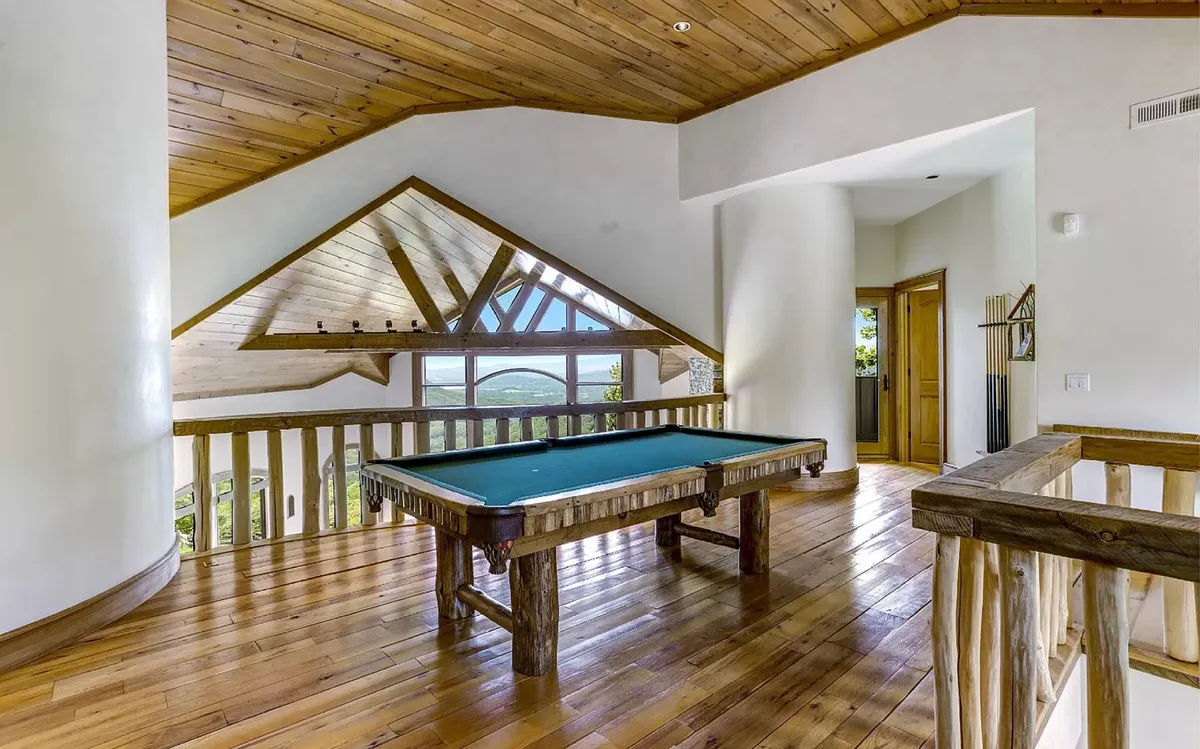 This image presents the loft’s game area from an alternate perspective, focusing on the pool table, expansive windows with scenic views, a wood-paneled ceiling, and the open feel of the railing. The setup conveys a relaxed, social environment ideal for gatherings and group activities.