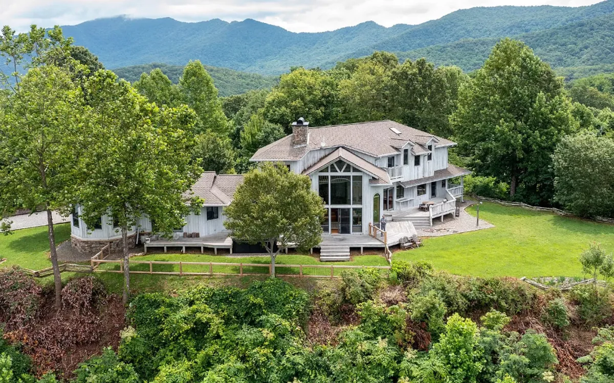 The image showcases a spacious, luxurious house with expansive windows set amid lush trees, all framed by breathtaking mountain vistas. The setting sun casts a golden glow over the property, hinting at the tranquil sunset views guests will enjoy from the porch. The serene landscape invites relaxation, outdoor gatherings, and exploration of the surrounding nature.