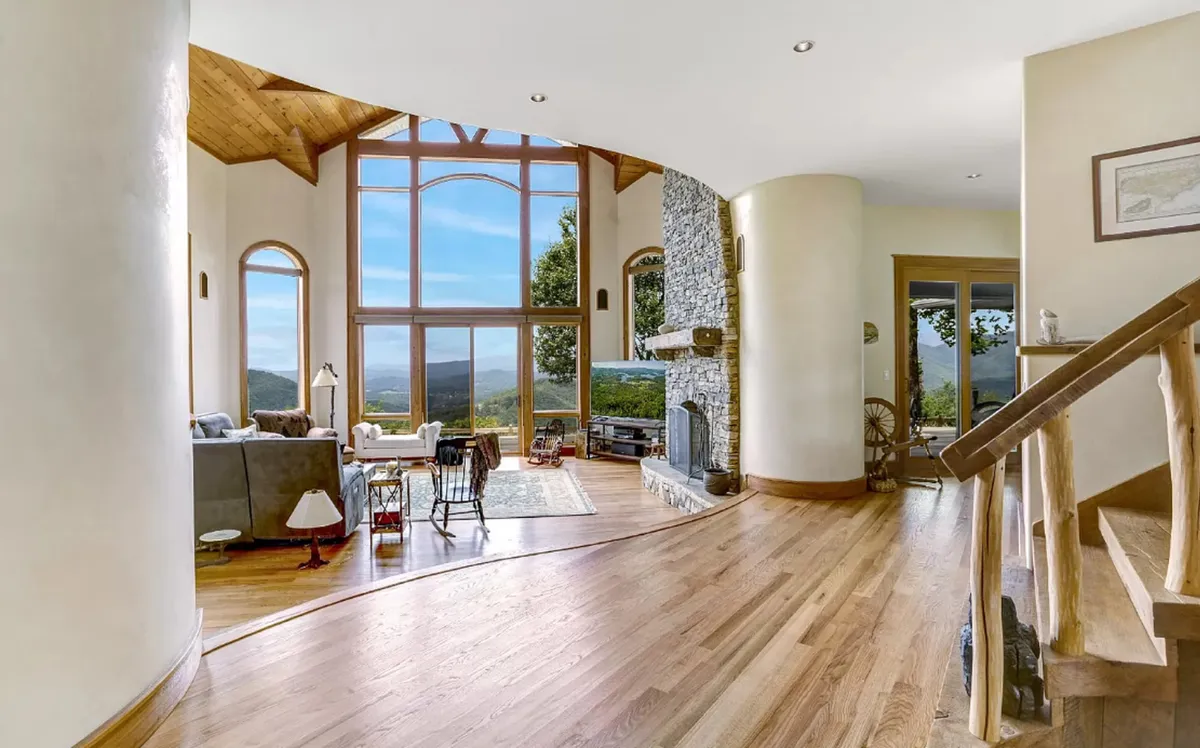 A grand living room with floor-to-ceiling windows that flood the space with natural light, framing stunning mountain views. A striking stone fireplace anchors the room, while curved walls and plush seating invite relaxation and social gatherings. The overall ambiance is both luxurious and welcoming, perfectly suited to memorable group experiences.