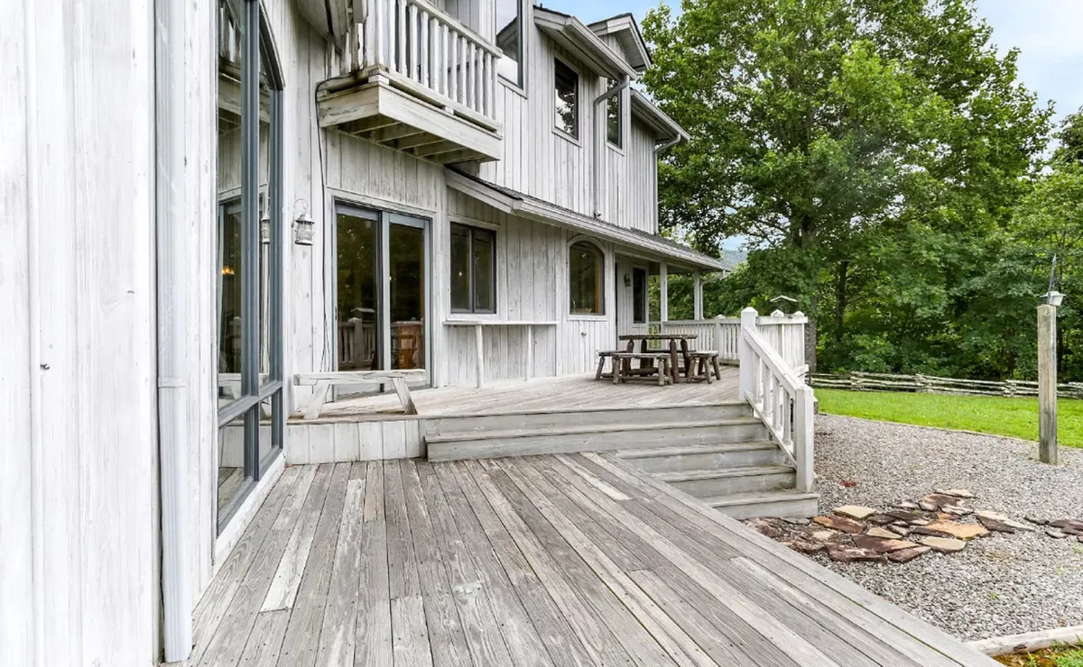 A spacious wooden deck furnished with a dining table, surrounded by a green lawn and mature trees. The area looks serene and is clearly designed for outdoor family gatherings and relaxation, with enough space for group meals or morning coffee in the fresh air.