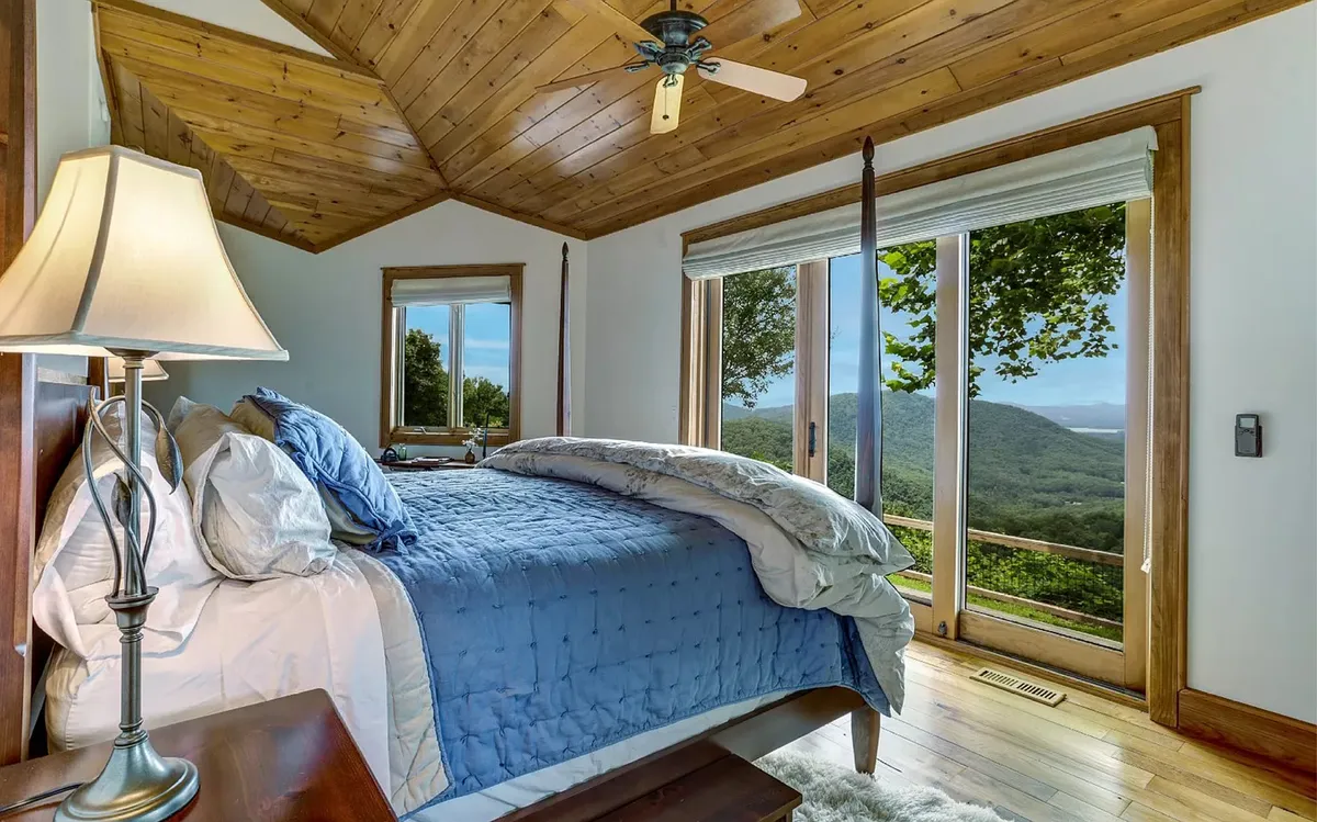 A serene bedroom featuring a king-size bed, large windows that fill the space with natural light, a cozy wooden ceiling, and a nightstand with a lamp. The tranquil setting invites relaxation and offers views that connect guests to the surrounding landscape.