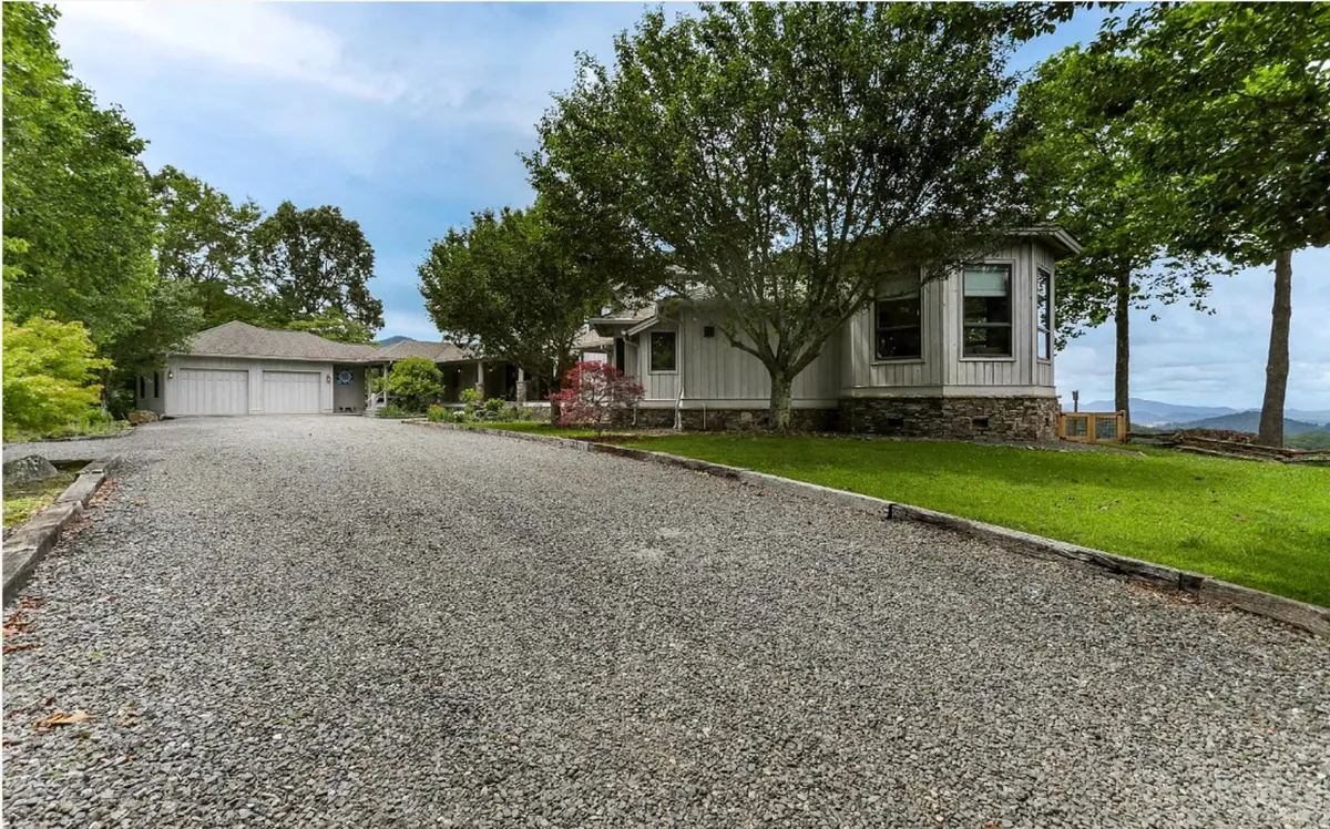 This image shows the tranquil exterior of the property, featuring a gravel driveway, the house itself nestled among mature trees, and a scenic mountain backdrop. The space suggests ample room for outdoor activities and the serenity of a natural setting.