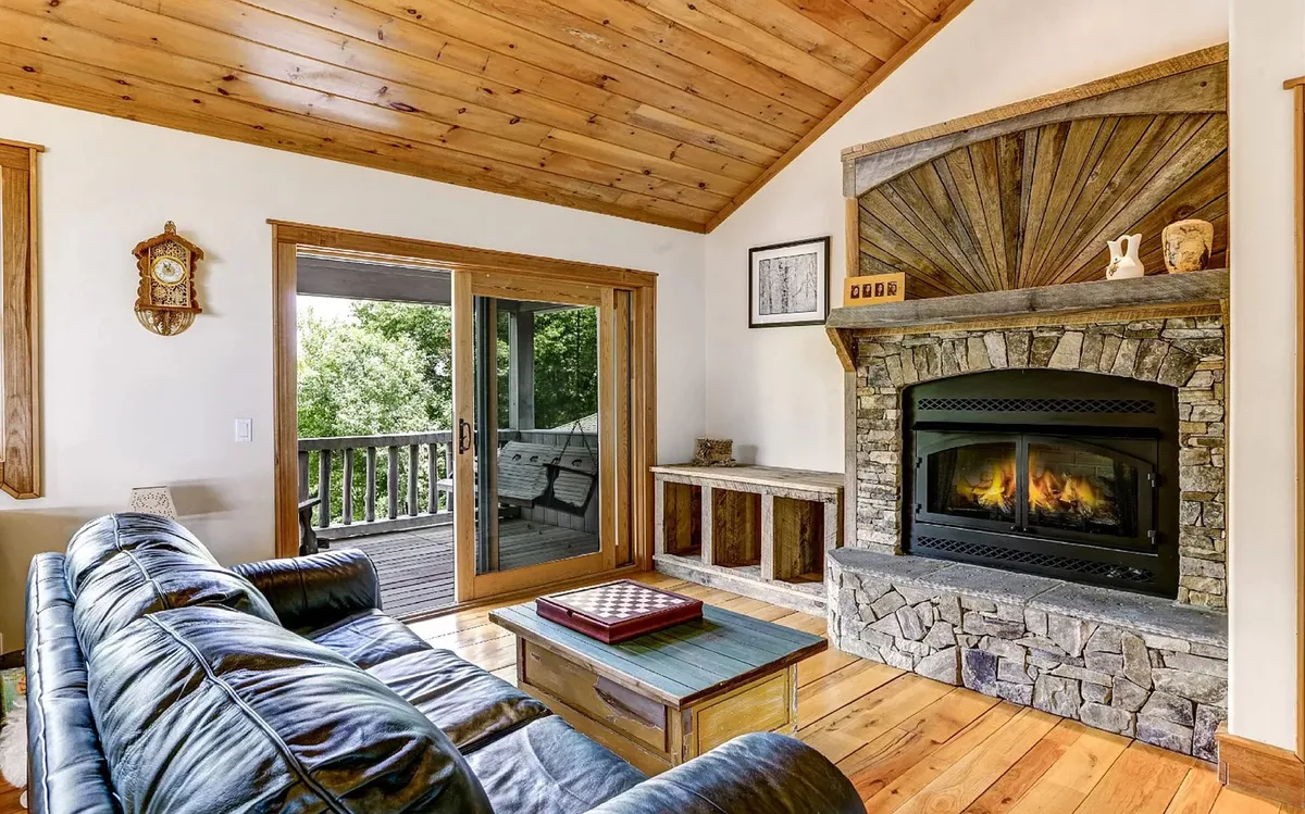 A warm and welcoming living area with a stone fireplace, wood-paneled ceiling, and floor-to-ceiling windows, offering panoramic views of the surrounding mountains. The space exudes comfort while keeping the natural environment front and center.