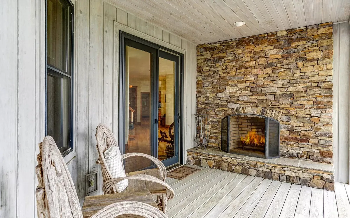 This image features a beautiful stone fireplace set on a spacious outdoor deck, flanked by inviting wooden rocking chairs and large glass doors that connect the inside to the outside. The setting exudes warmth and relaxation, perfect for enjoying a drink, reading, or gathering with others as dusk settles over the mountain landscape.
